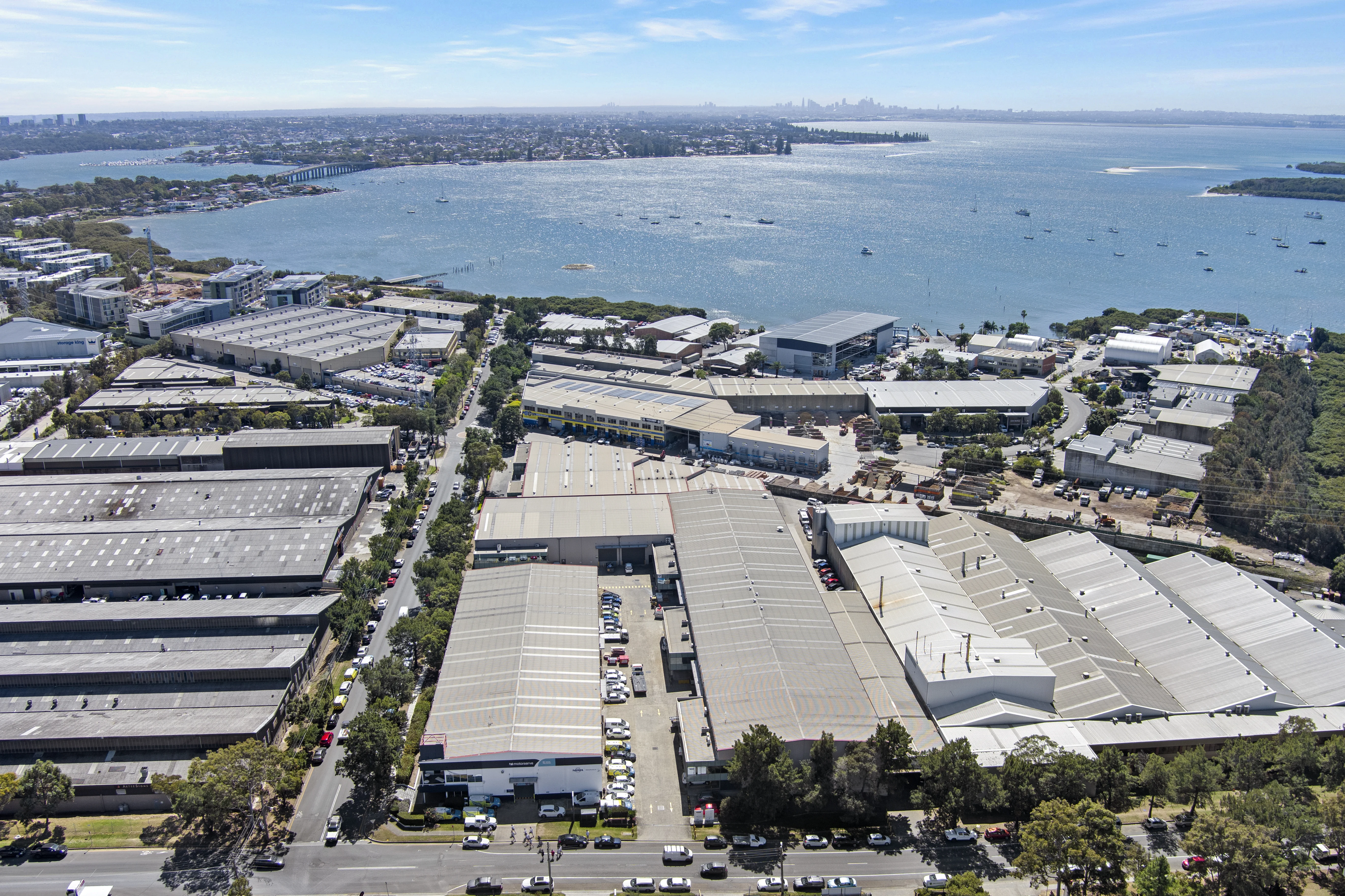 Aerial view of an industrial warehouse complex near a waterfront, with a city skyline in the background.